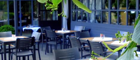 Zona pranzo all'aperto con tavoli e sedie, circondata da ampie finestre.