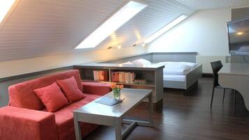 Ein modernes Zimmer mit schrägem Dach, einem roten Sofa, einem kleinen Tisch mit einem Buch, einem Bett, einem Fernseher und einem Bücherregal.