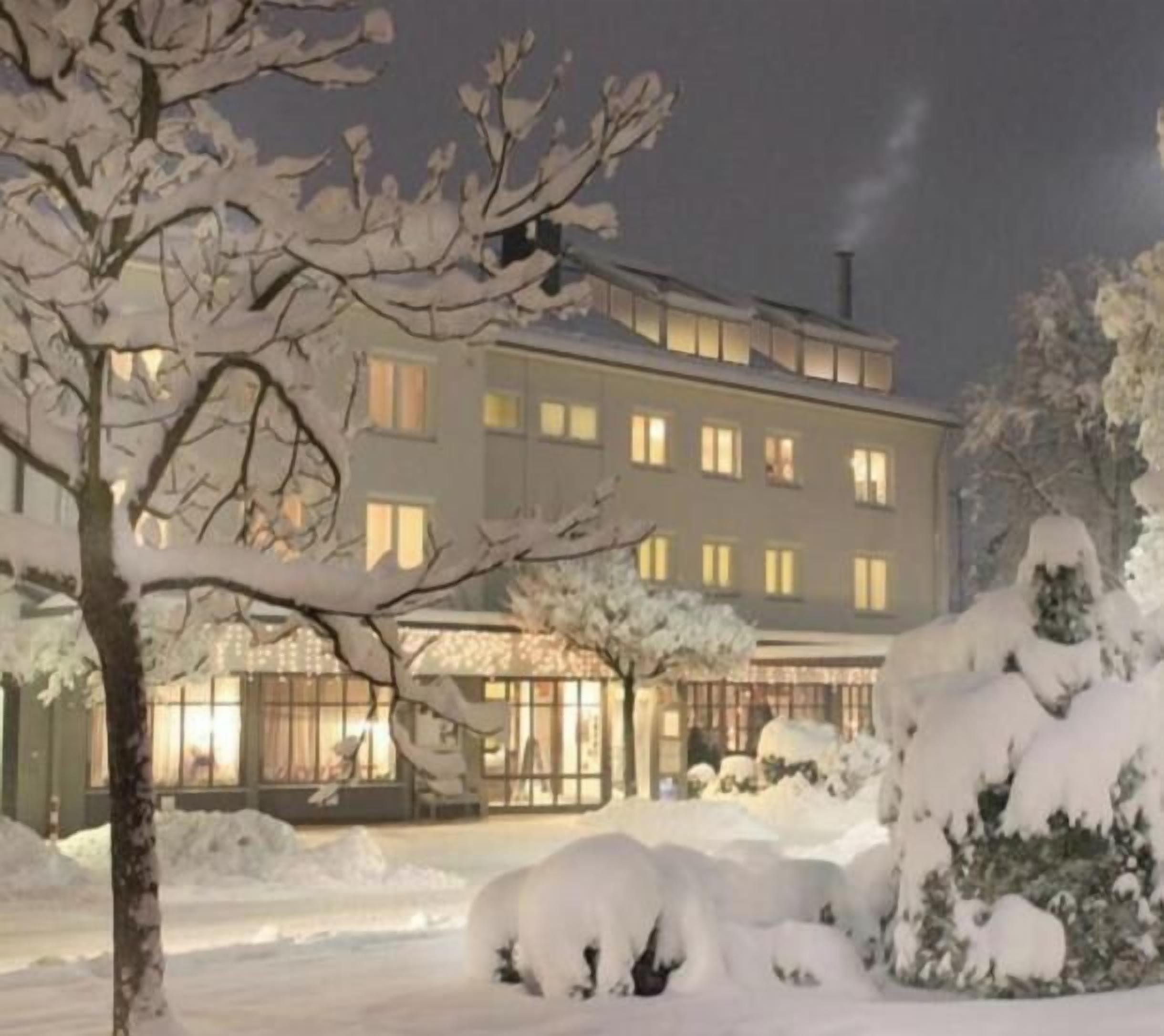 Ein mehrstöckiges Gebäude mit verschneiter Landschaft, beleuchteten Fenstern und einem gut sichtbaren Eingang.