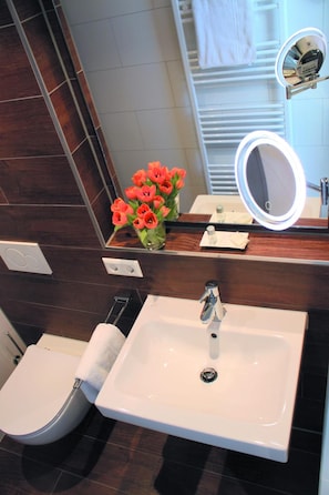 Un baño con lavamanos blanco, inodoro, bidé, espejo con luz y un jarrón con flores.