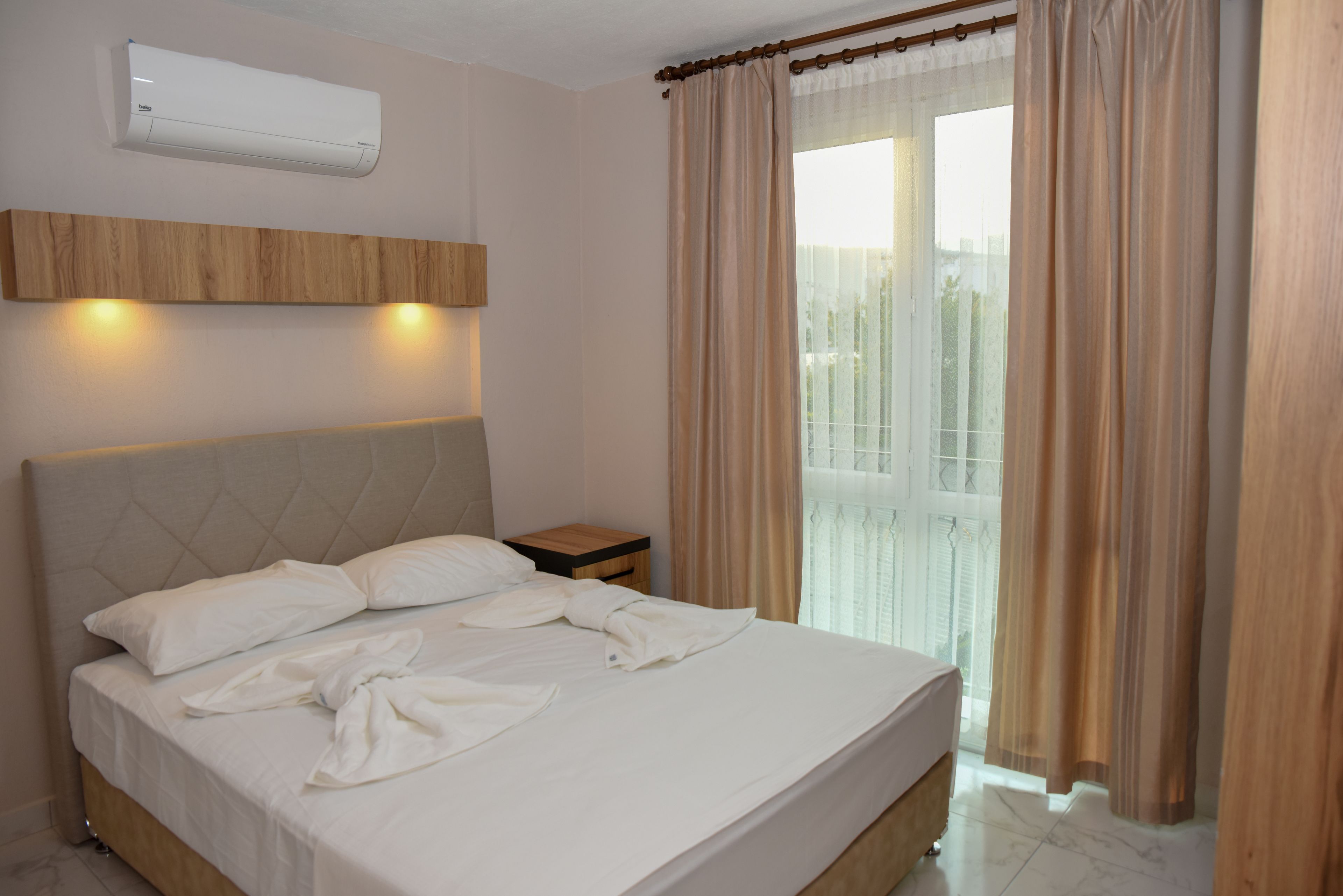 A neatly made bed with white linens, a wooden headboard, a bedside table, and an air conditioning unit mounted on the wall.