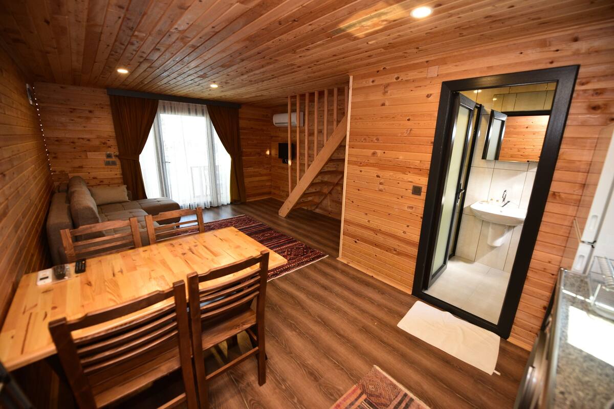 A wooden cabin interior with a dining area, sofa, staircase, and a bathroom visible through a glass door.