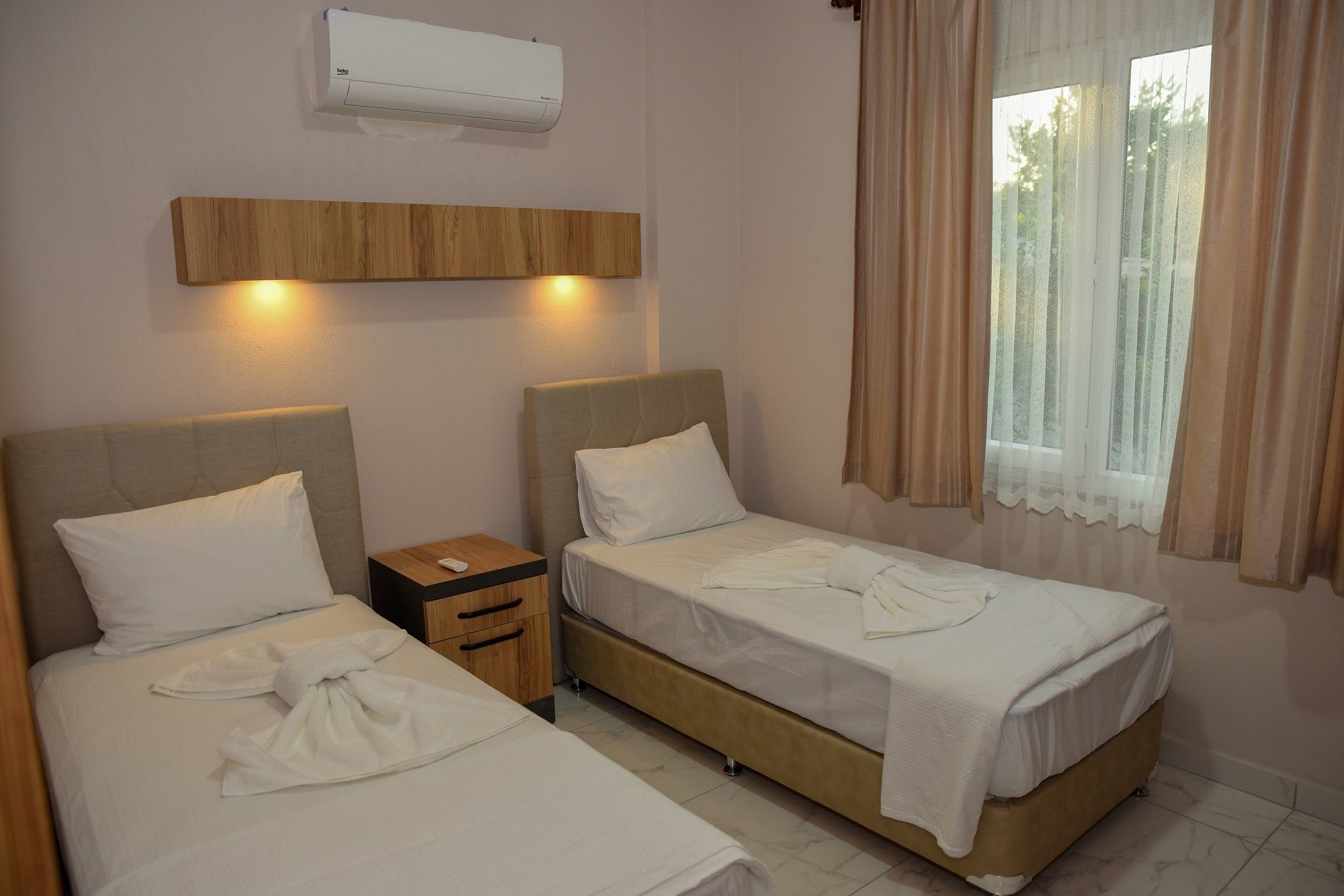 A hotel room with two single beds, a bedside table with a remote control, an air conditioning unit, and a window with curtains.