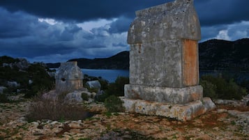 A stone monument with a weathered appearance, surrounded by rocky terrain and vegetation, with a body of water and mountains in the background.