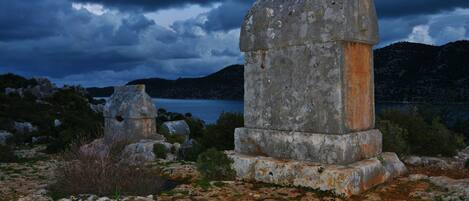 Ein steinernes Monument mit verwitterter Oberfläche, umgeben von felsigem Gelände und Vegetation, mit einem Gewässer und Bergen im Hintergrund.