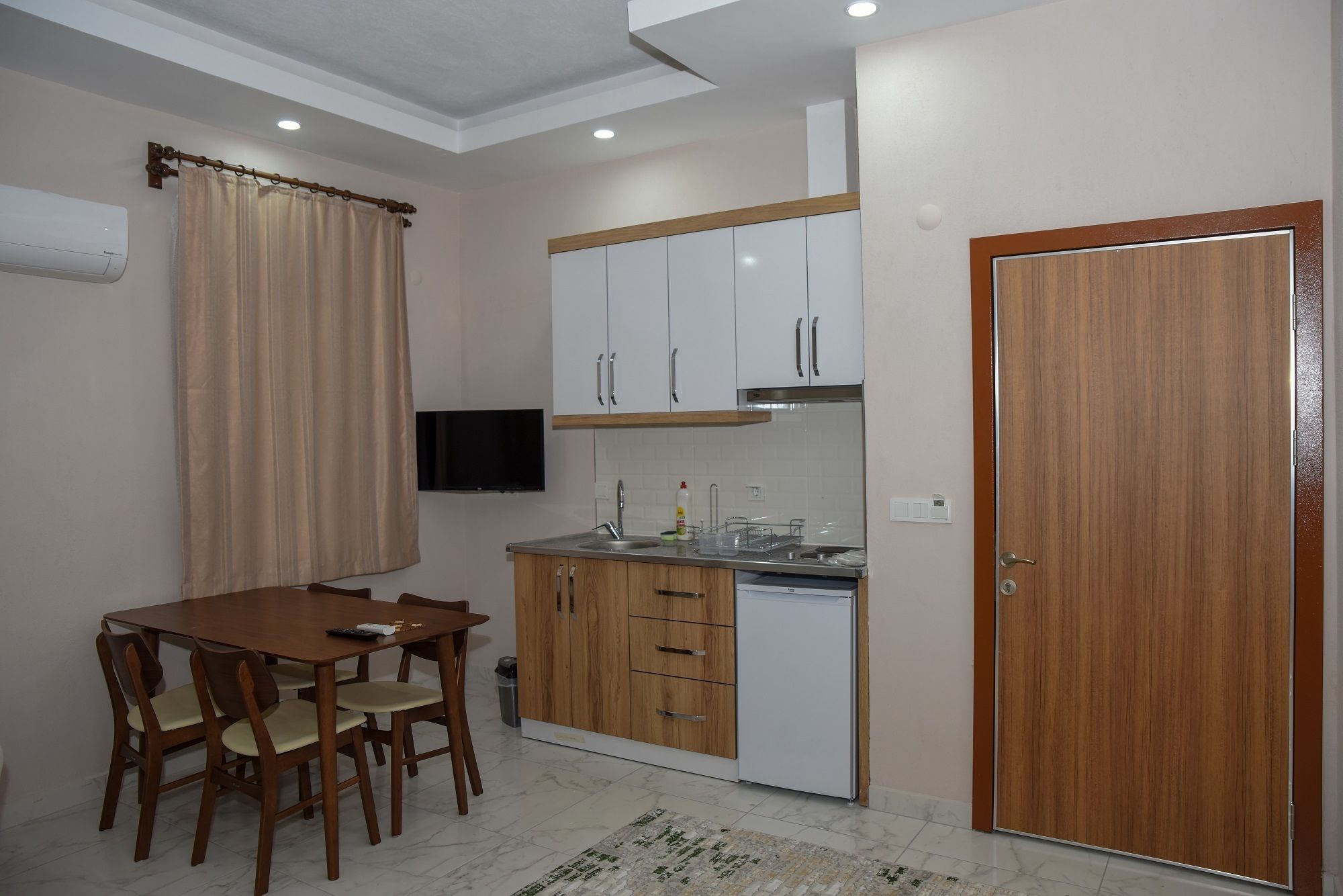 A compact kitchen with white cabinets, a wooden dining table, and a TV mounted on the wall.