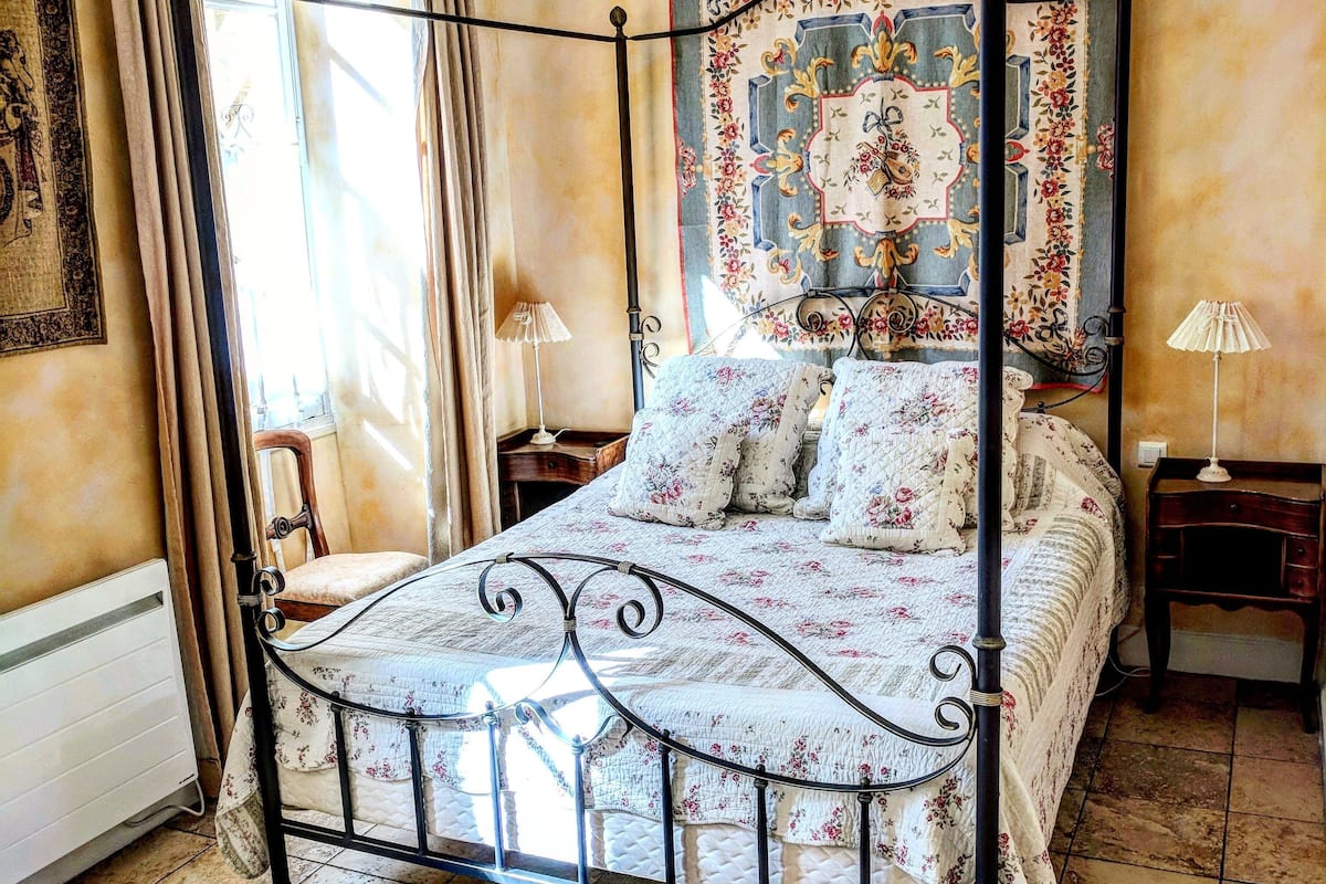 Un lit à baldaquin avec une couvre-lit à motifs floraux, une tapisserie au mur et une vue sur l’extérieur par une fenêtre ornée de rideaux.