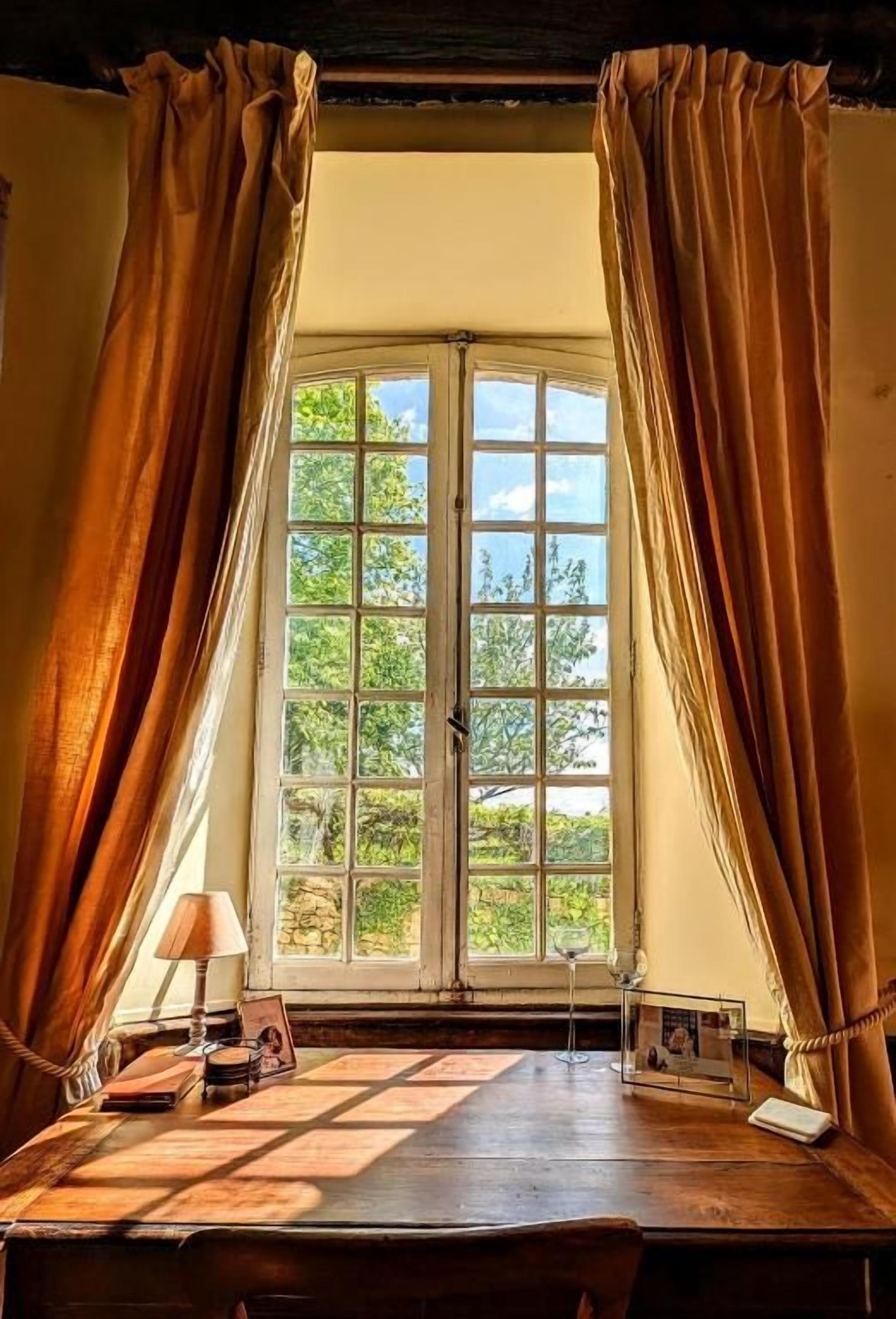 A room with a large window, curtains, a wooden table, and a lamp.