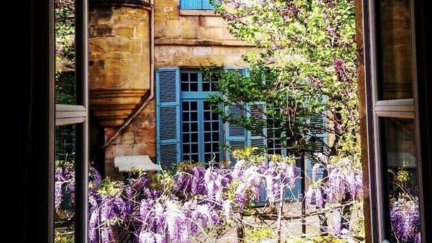 Un edificio de piedra con contraventanas azules y un jardín con flores púrpuras.