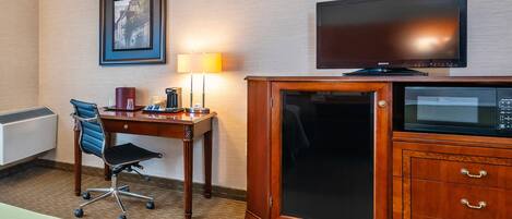 Quarto de hotel com uma cama, uma escrivaninha com luminária, uma televisão e um armário.