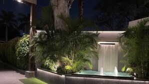 Espacio exterior iluminado por la noche, con fuente de agua, plantas iluminadas y un camino pavimentado.