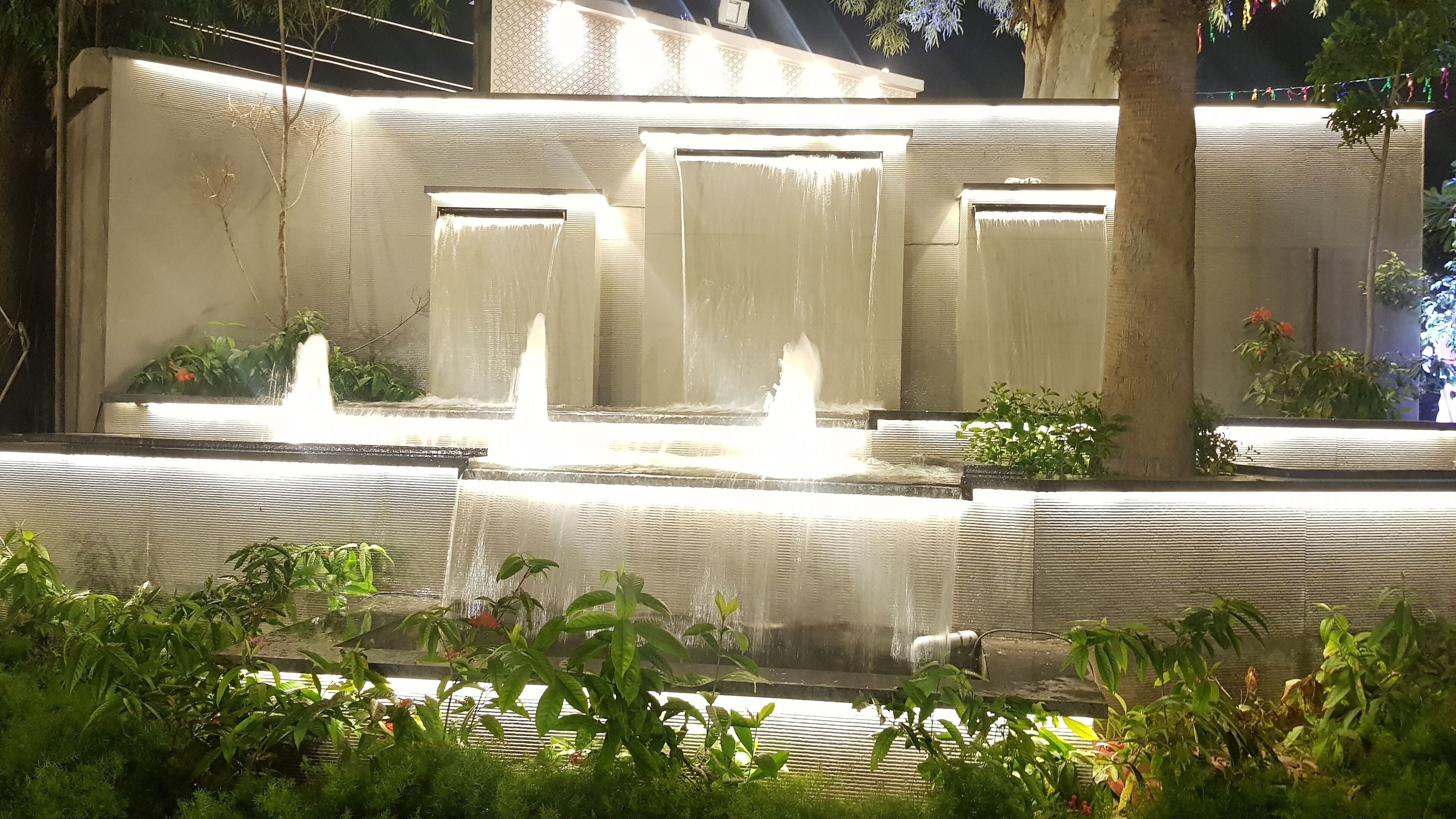 A nighttime fountain with multiple water jets, surrounded by greenery and a building with decorative lighting.