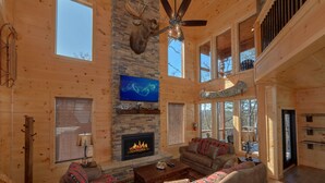 A spacious living room with a stone fireplace, a ceiling fan, and a large TV mounted on the wall. - Family Pool Cabin, Theater, Gameroom, Hot Tub (Gatlinburg)
