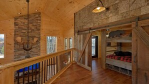 A wooden cabin interior with a stone fireplace, a wooden staircase, and a loft bed with a bear decoration. - Family Pool Cabin, Theater, Gameroom, Hot Tub (Gatlinburg)