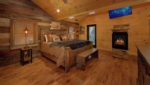 A cozy log cabin bedroom with a bed, a bench, a fireplace, and a ceiling fan. - Family Pool Cabin, Theater, Gameroom, Hot Tub (Gatlinburg)