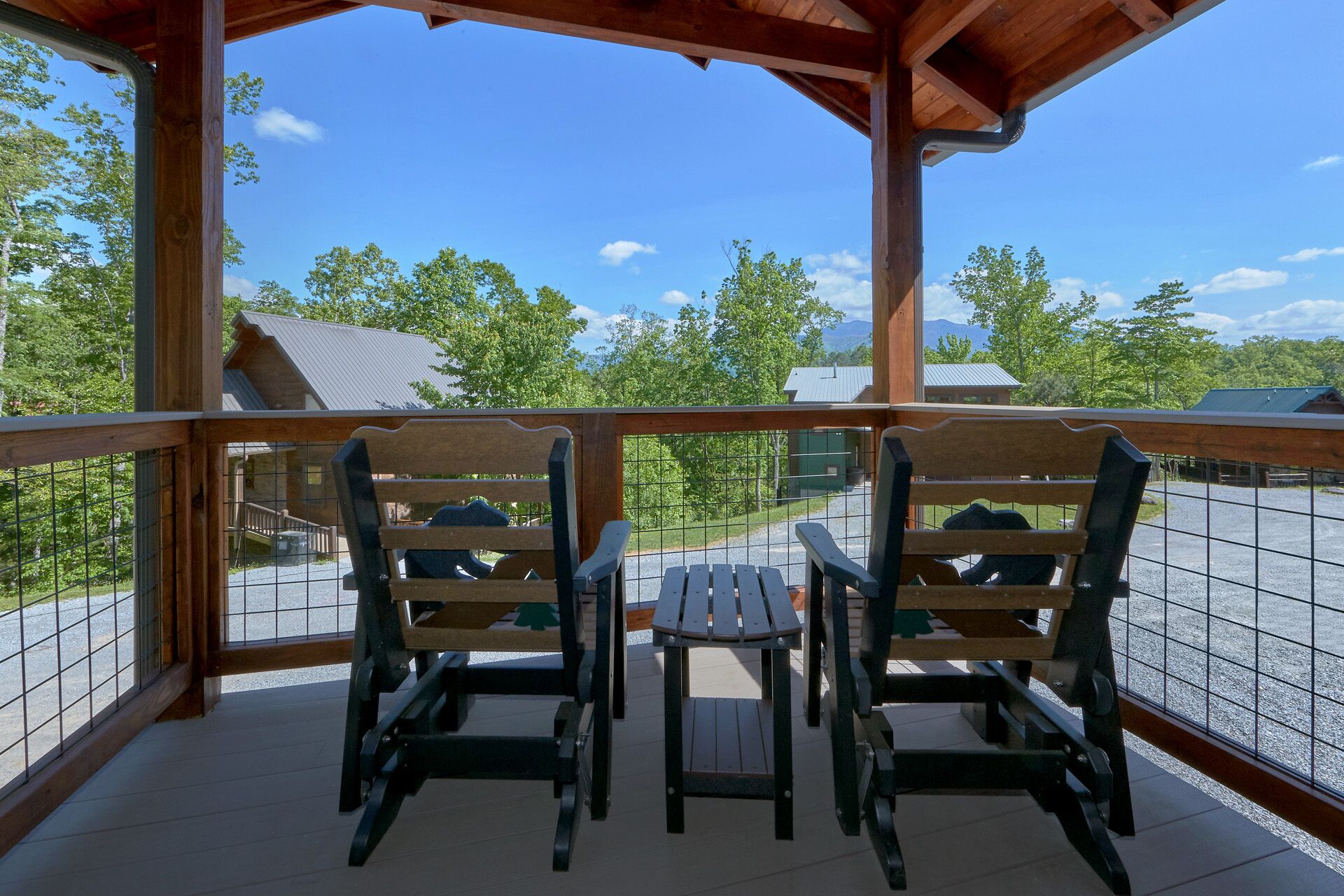 Un espacio al aire libre cubierto con dos sillas Adirondack y una mesa pequeña, con vista a un estacionamiento y edificios en un área boscosa.
