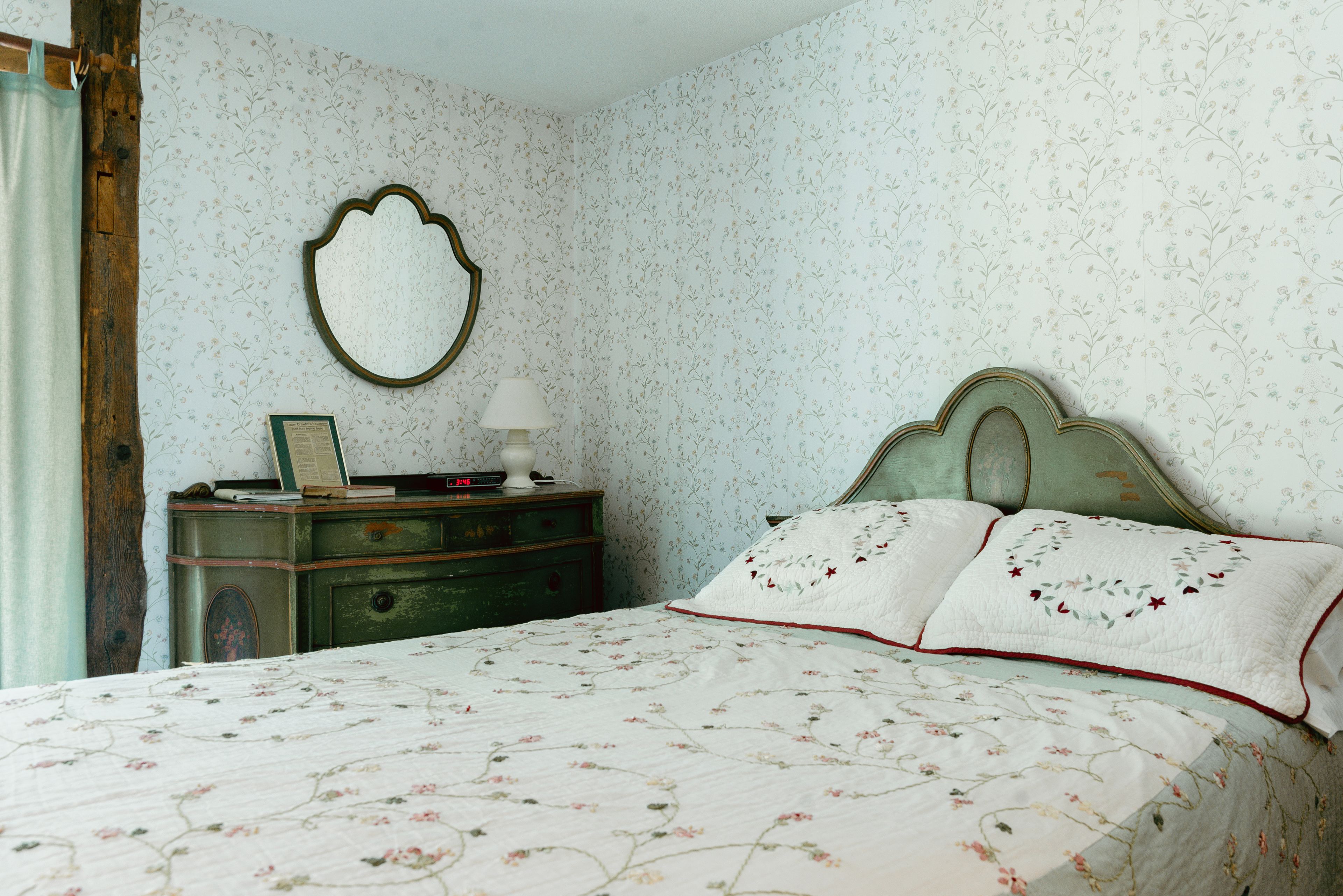 A bedroom with floral wallpaper, a vintage dresser, a bed with floral bedding, and a mirror on the wall.