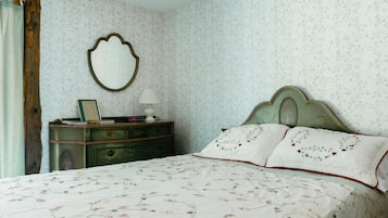 Un dormitorio con papel tapiz floral, un tocador antiguo, una cama con ropa de cama floral y un espejo en la pared.