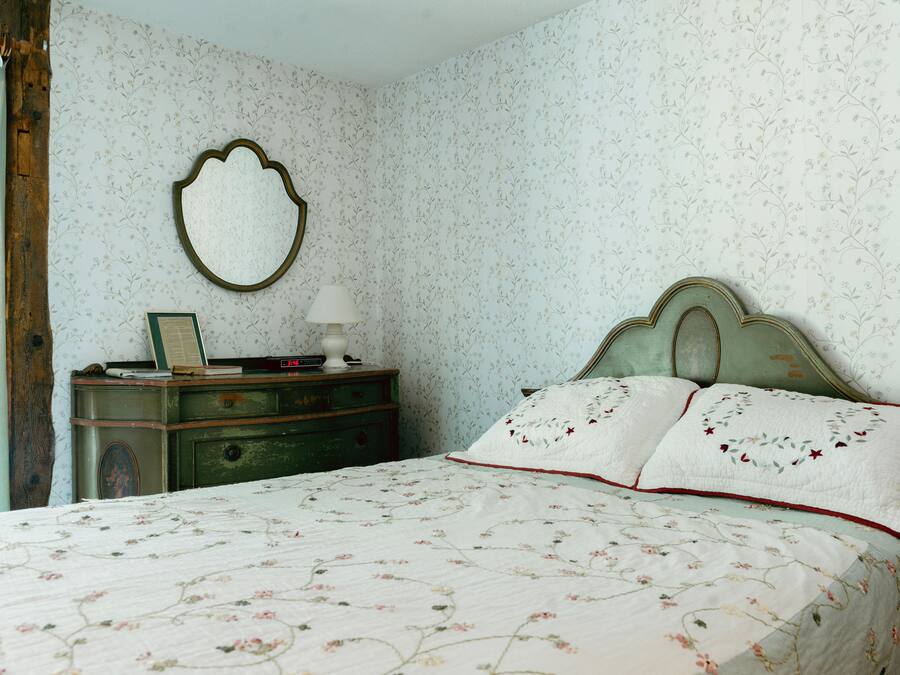 A bedroom with floral wallpaper, a vintage dresser, a bed with floral bedding, and a mirror on the wall.