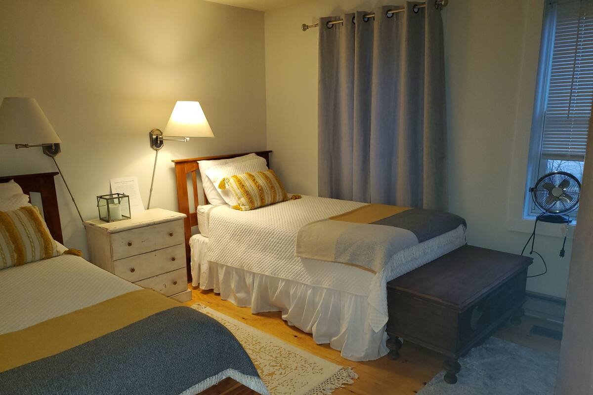 Une chambre avec deux lits, une table de chevet, une lampe, un ventilateur et une fenêtre avec des stores.