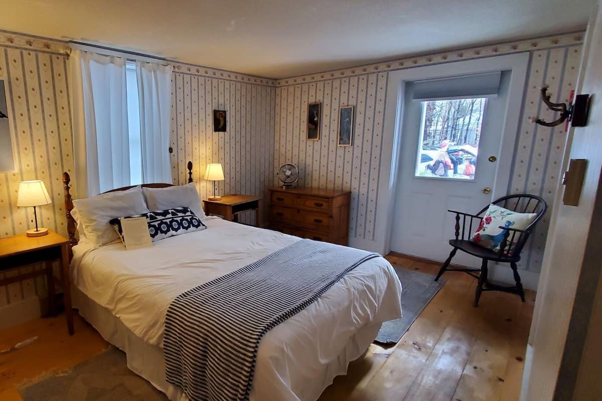 Une chambre à coucher comprenant un lit, deux tables de chevet avec des lampes, une chaise, une commode et une porte donnant sur un couloir.