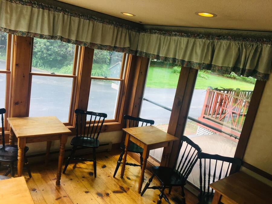 A corner room with wooden flooring, a wooden table, and four chairs. Large windows with curtains are visible, offering a view of a green landscape and a red deck outside.