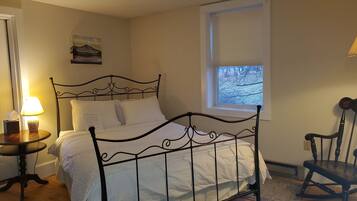 A neatly made bed with a metal frame, a bedside table with a lamp, a chair, and a window with a view of trees.