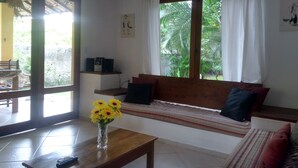 Ein Zimmer mit einer hölzernen Bank, einem kleinen Tisch mit einer Vase voller Sonnenblumen, einem Fenster mit Vorhängen und Blick ins Grüne.