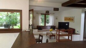 Ein Wohnzimmer mit Sofa, Fernseher und Couchtisch. Ein Fenster bietet Blick auf Grünflächen.