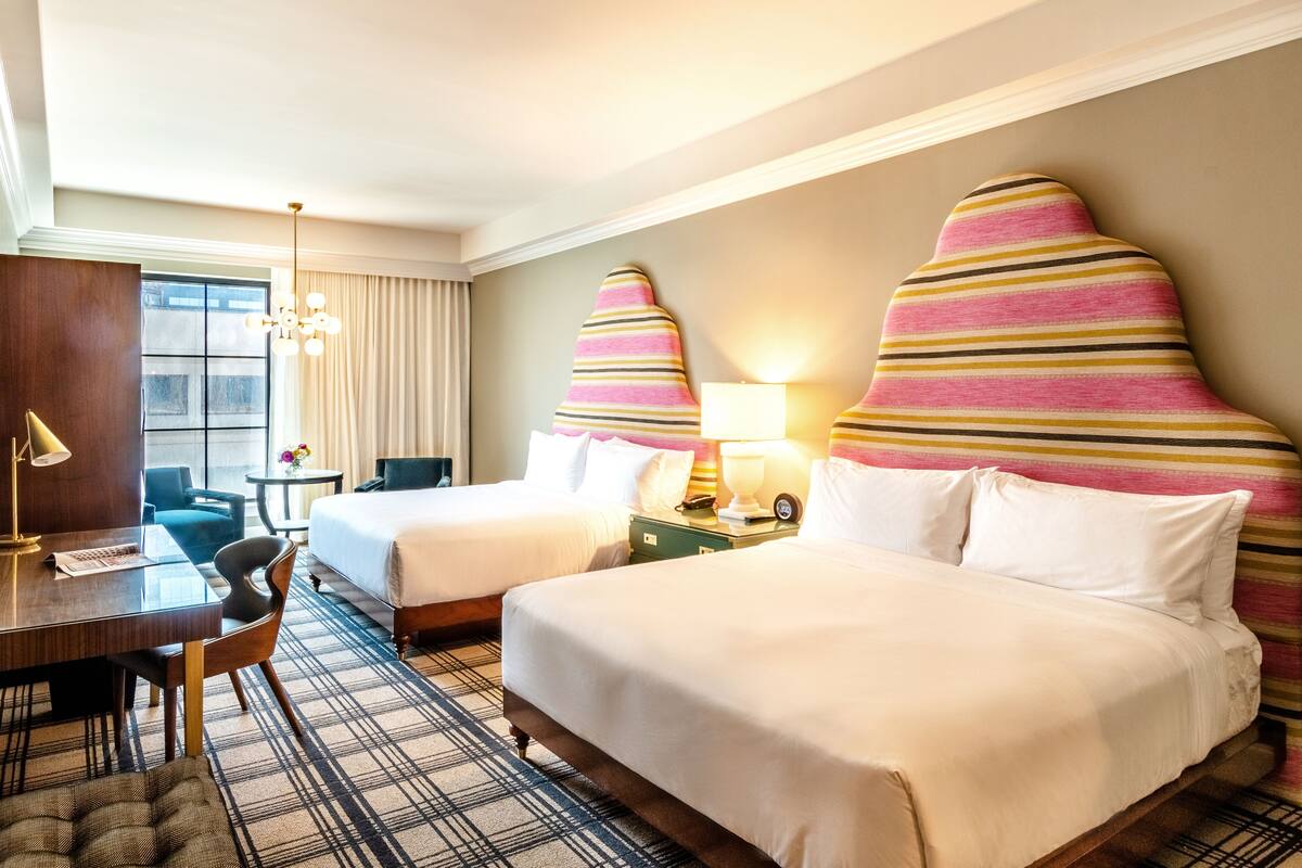 A hotel room with two beds, a desk, a chair, a lamp, and a window with curtains.