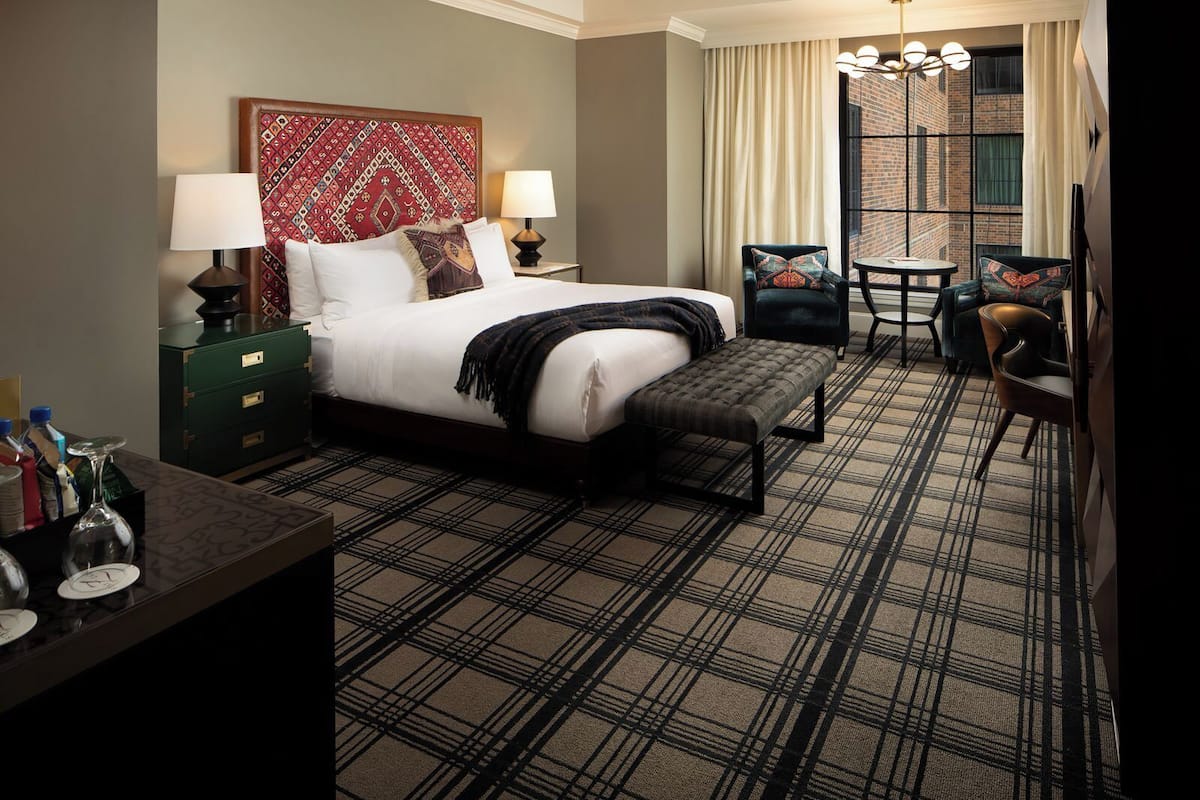 A hotel room with a large bed, two bedside tables, a desk, a chair, and a view of a brick building through a window.