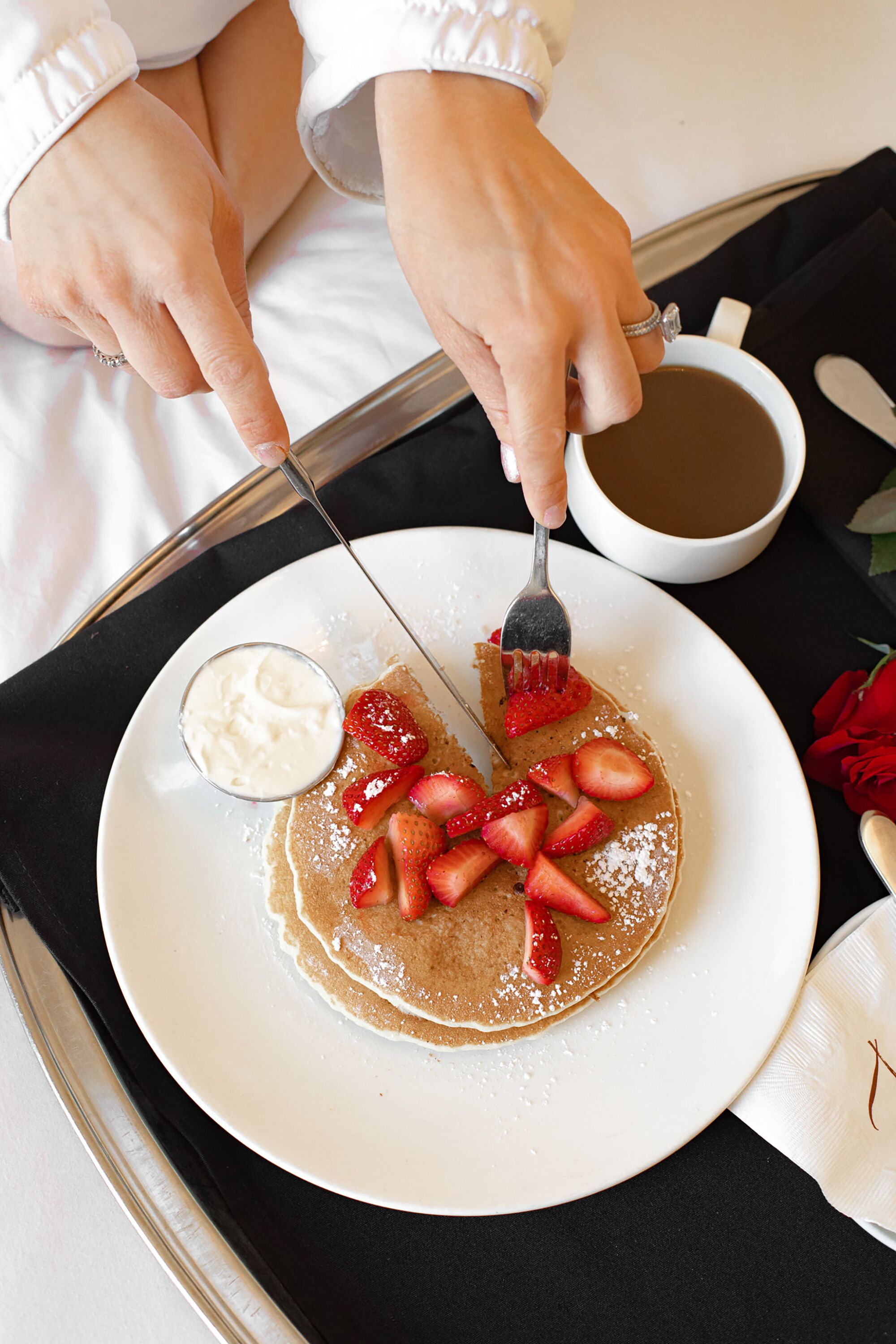 A plate of pancakes with strawberries and whipped cream, a cup of coffee, and a rose.