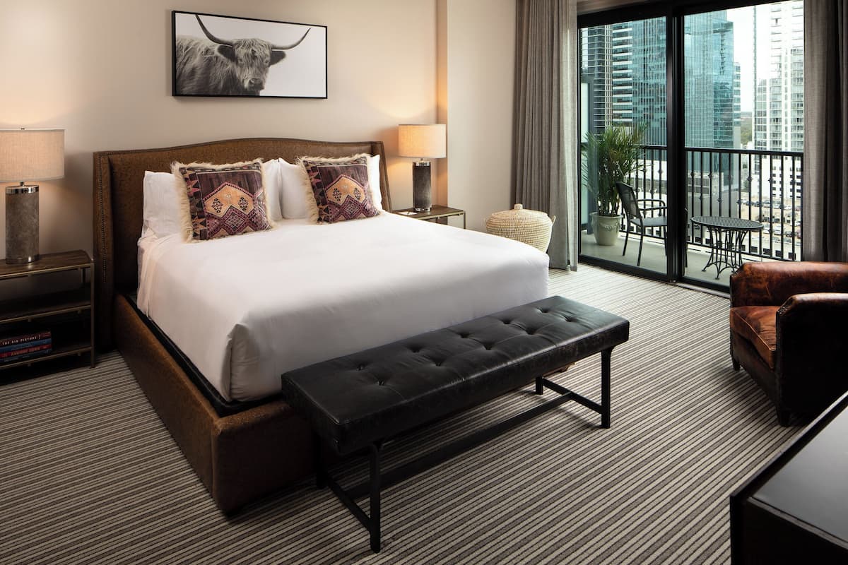 A hotel room with a large bed, a bench, a chair, a small table, a lamp, and a view of the city through a balcony.