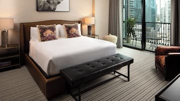 Une chambre d’hôtel avec un grand lit, une banquette, une chaise, une petite table, une lampe et une vue sur la ville depuis un balcon.