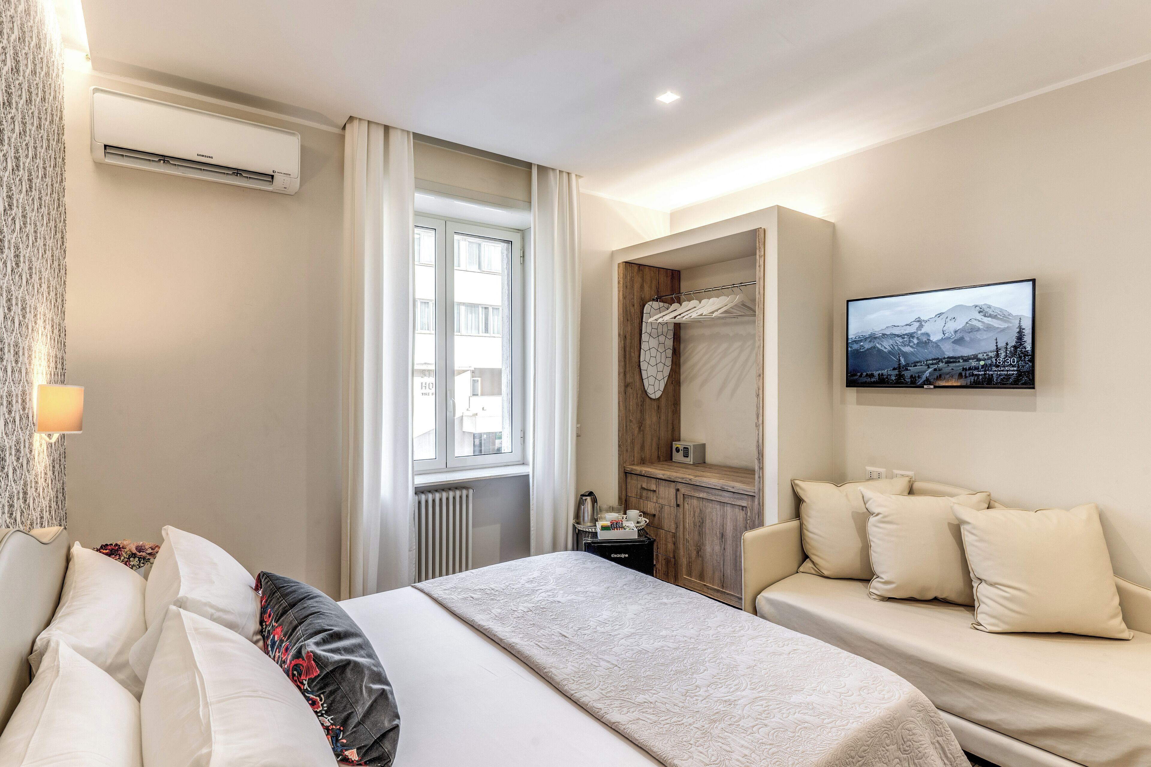 A hotel room with a bed, a sofa, an air conditioning unit, a television, and a window with curtains.