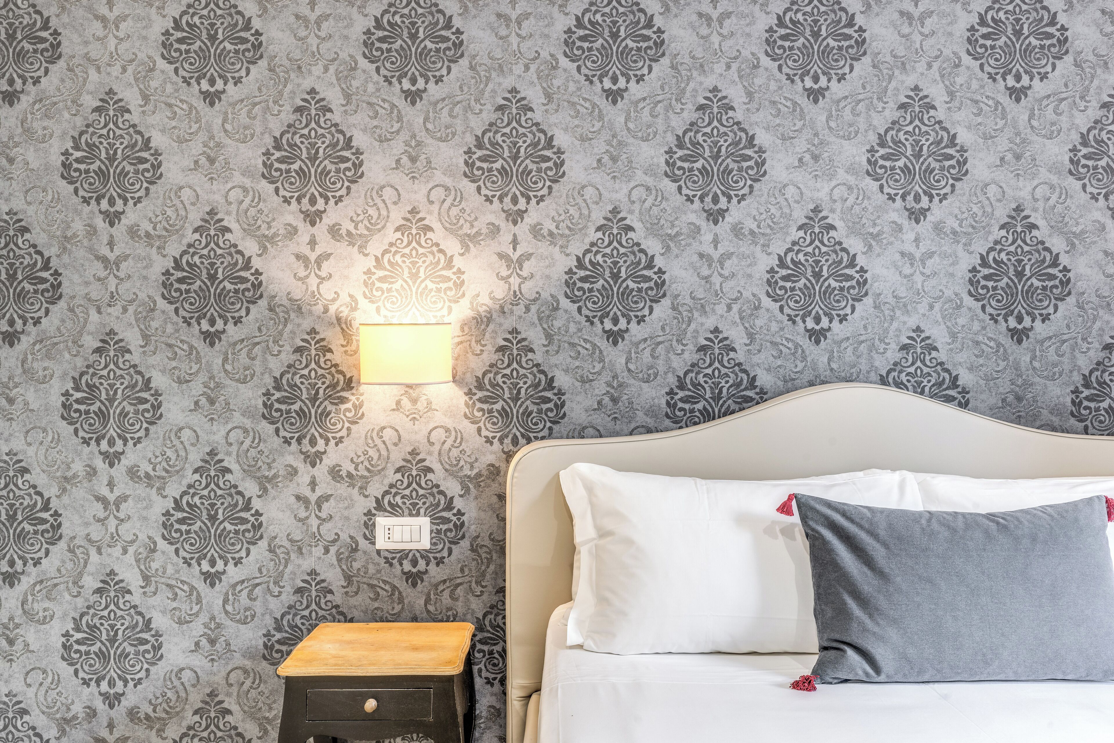 A neatly made bed with white and gray pillows, a bedside table with a lamp, and a patterned wallpaper.