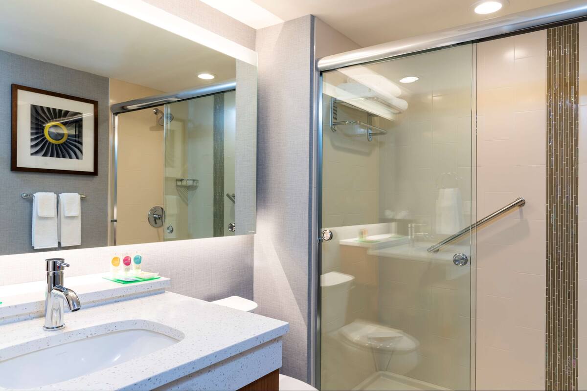 Un baño moderno con ducha envuelta en vidrio, un lavabo blanco con encimera moteada y una obra de arte enmarcada en la pared.