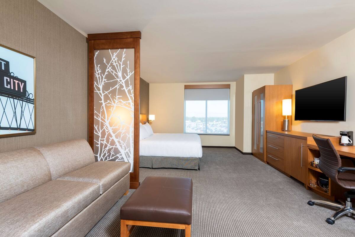 Habitación de hotel con cama, sofá, escritorio, televisión y ventana con vista a la ciudad.