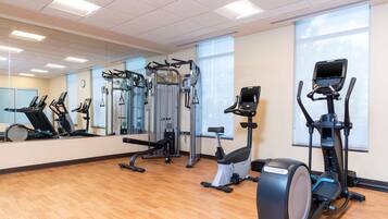 Een sportschool met diverse fitnessapparaten, grote ramen en een houten vloer.