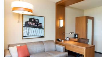 A hotel room with a grey sofa, a wooden desk, a mirror, a framed sign, a lamp, and a coffee maker.