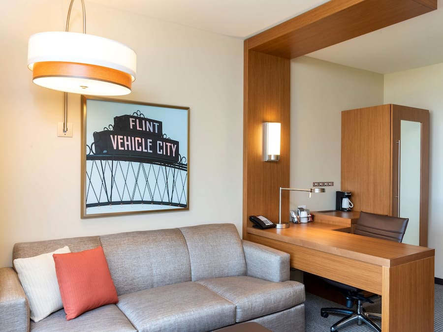 A hotel room with a grey sofa, a wooden desk, a mirror, a framed sign, a lamp, and a coffee maker.
