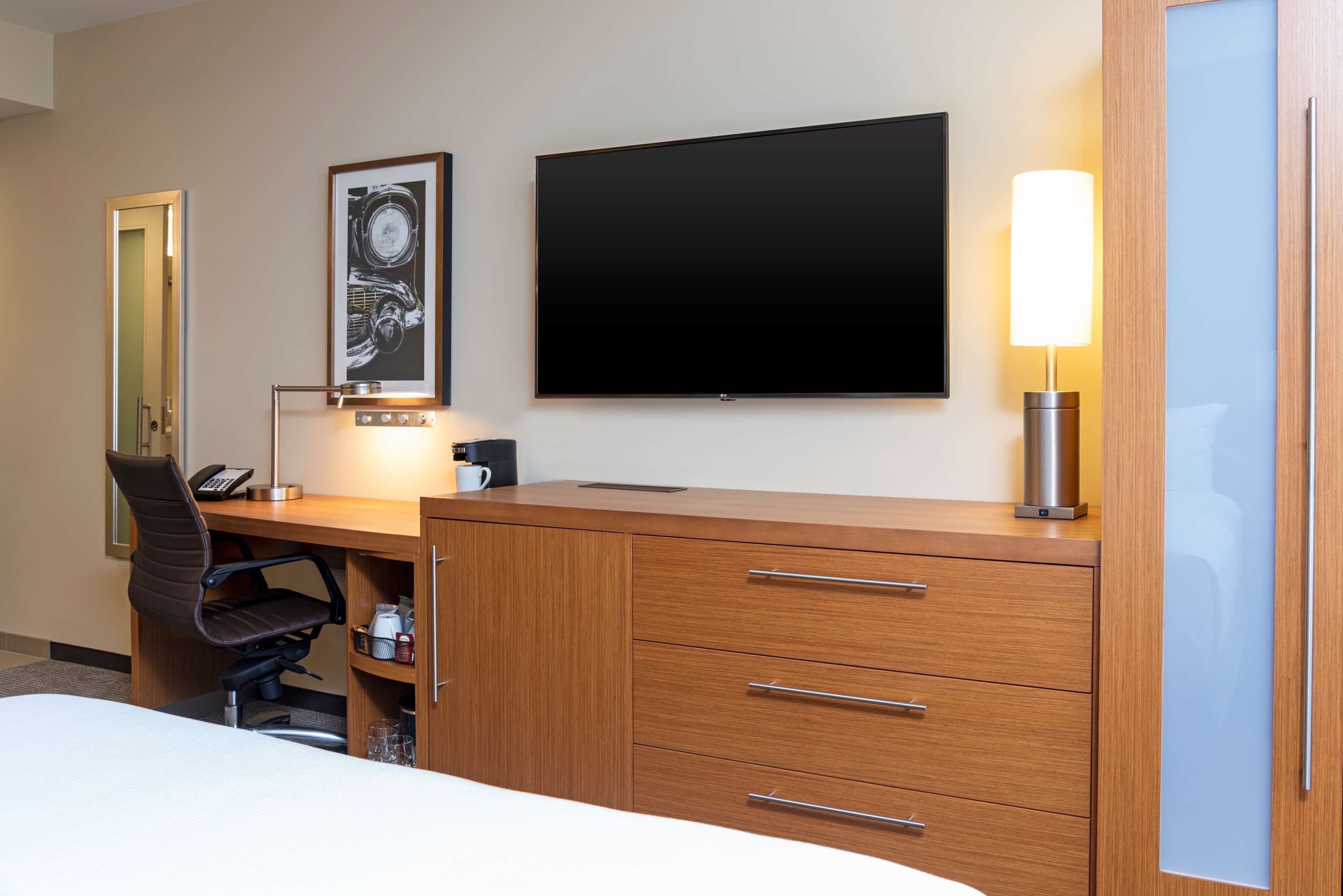 A hotel room with a bed, a desk with a chair, a flat-screen TV, a lamp, and a framed picture on the wall.