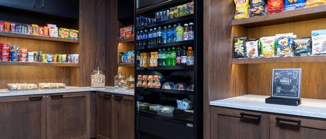 Una cocina con armarios de madera, un refrigerador lleno de bebidas y estantes con aperitivos y condimentos.