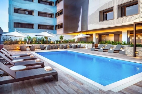 A modern hotel pool area with lounge chairs and umbrellas, surrounded by a building with a geometric facade.. Hyatt Place Aruba