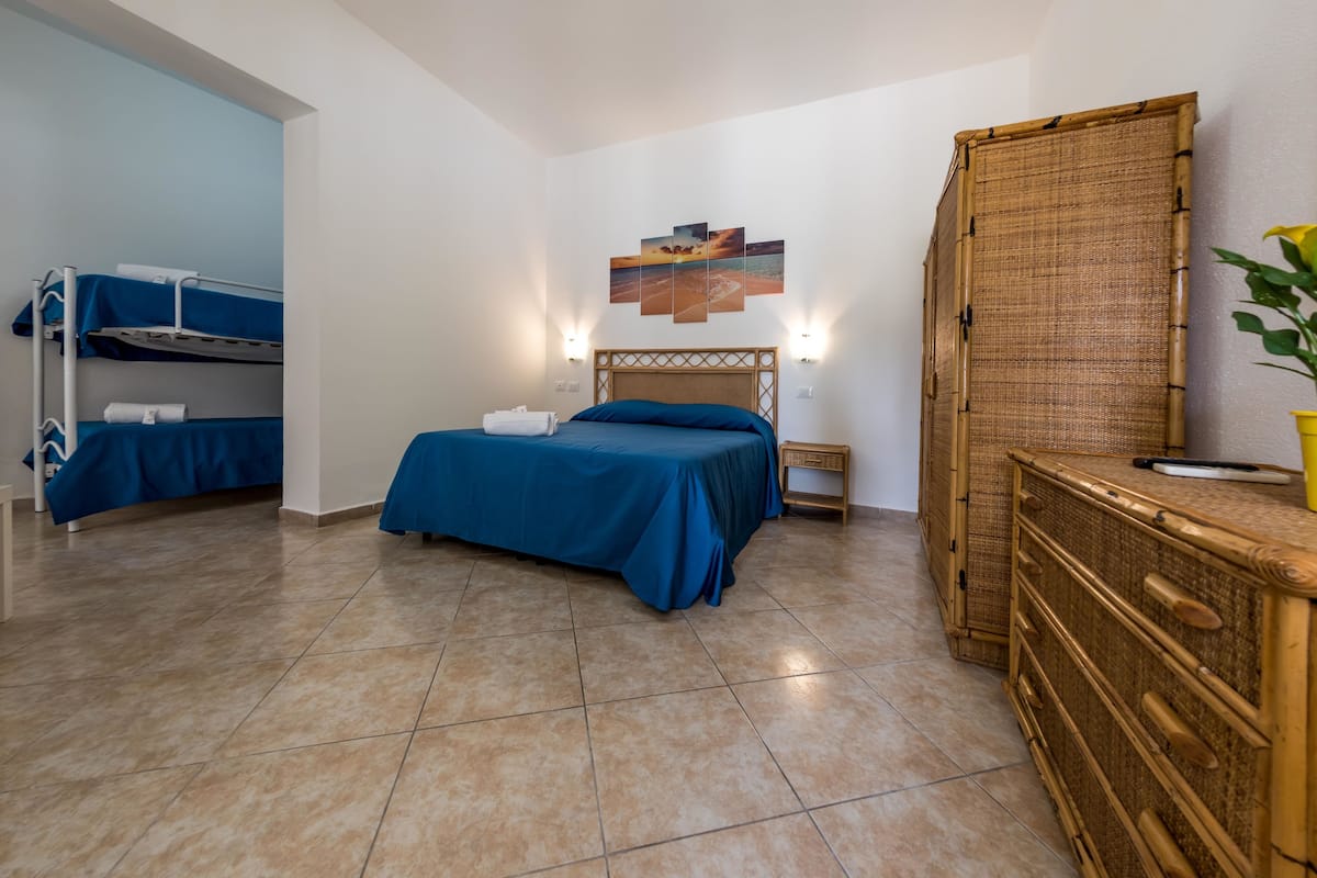 A hotel room with a bunk bed, a single bed with a blue bedspread, a wooden dresser, and a wall-mounted shelf.
