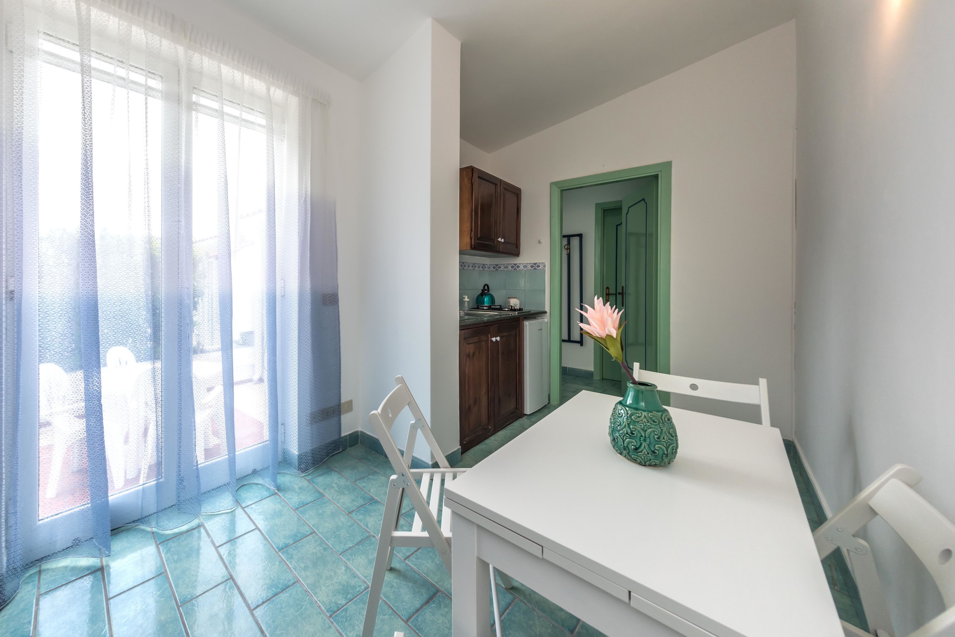 Un comedor con una mesa blanca, sillas y un florero con flores. Se ve una cocina al fondo con armarios de madera y una puerta verde.