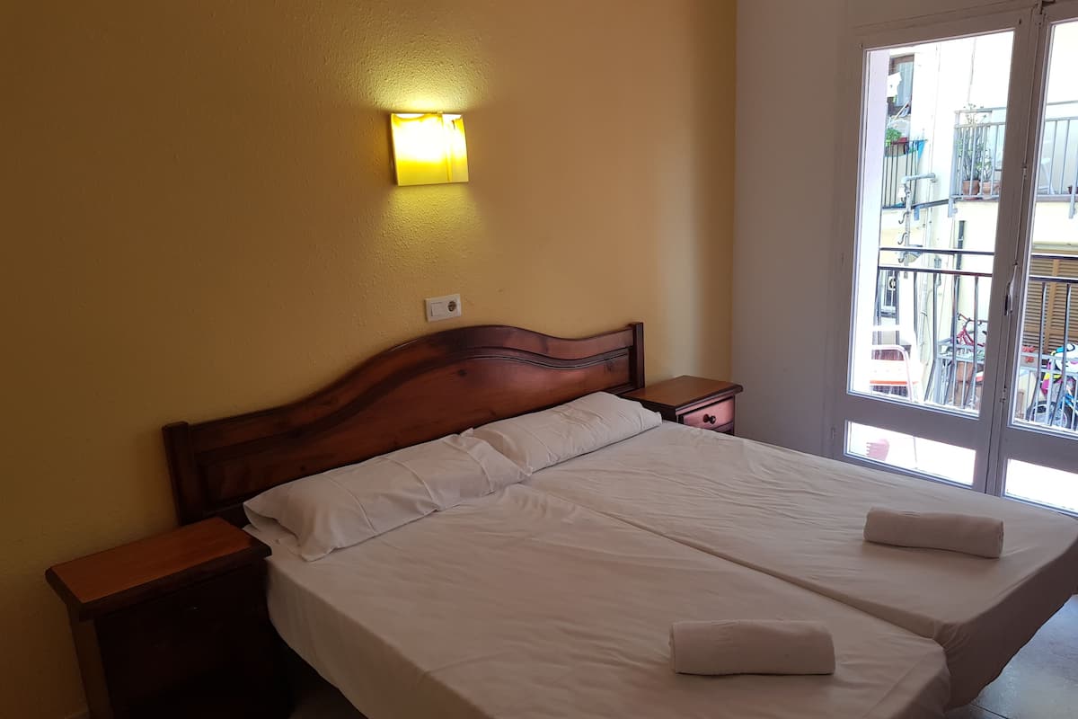 A double bed with white linens, a wooden headboard, a nightstand, a wall-mounted light, and a balcony view.