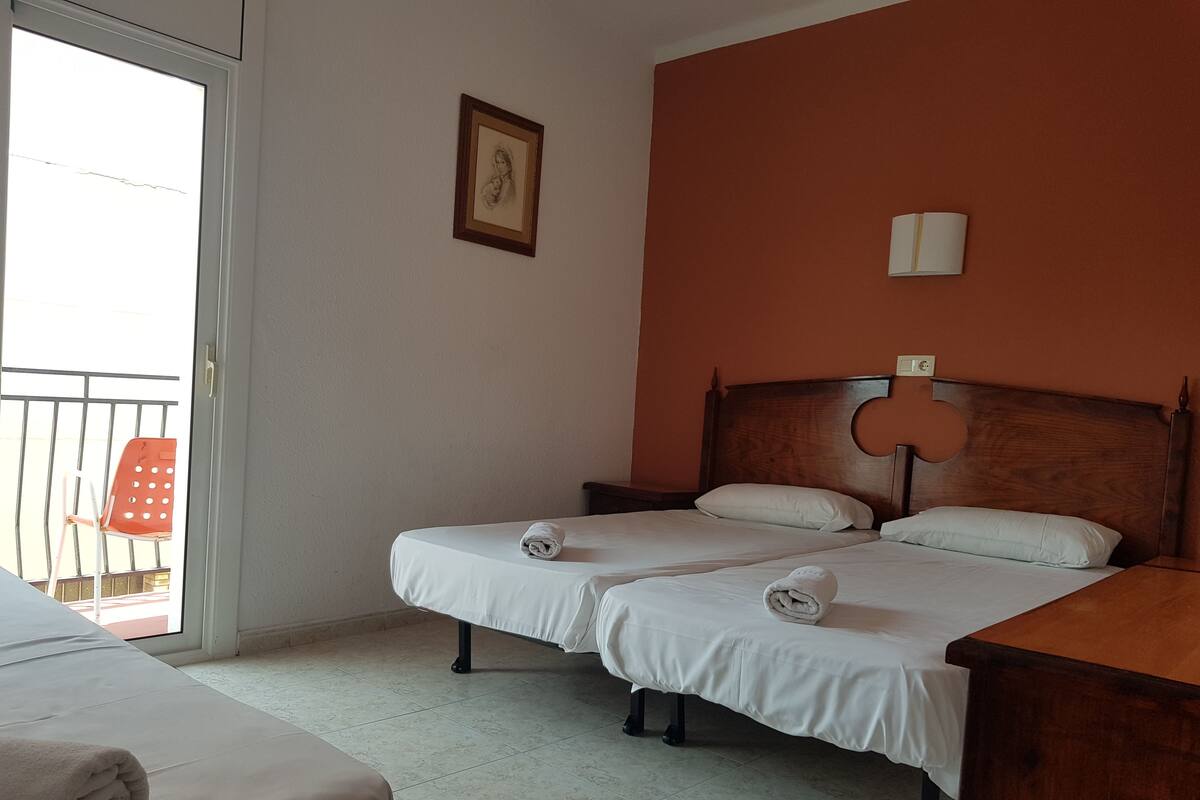 A hotel room with two beds, a wooden headboard, a small table, a chair, and a balcony with a red chair.