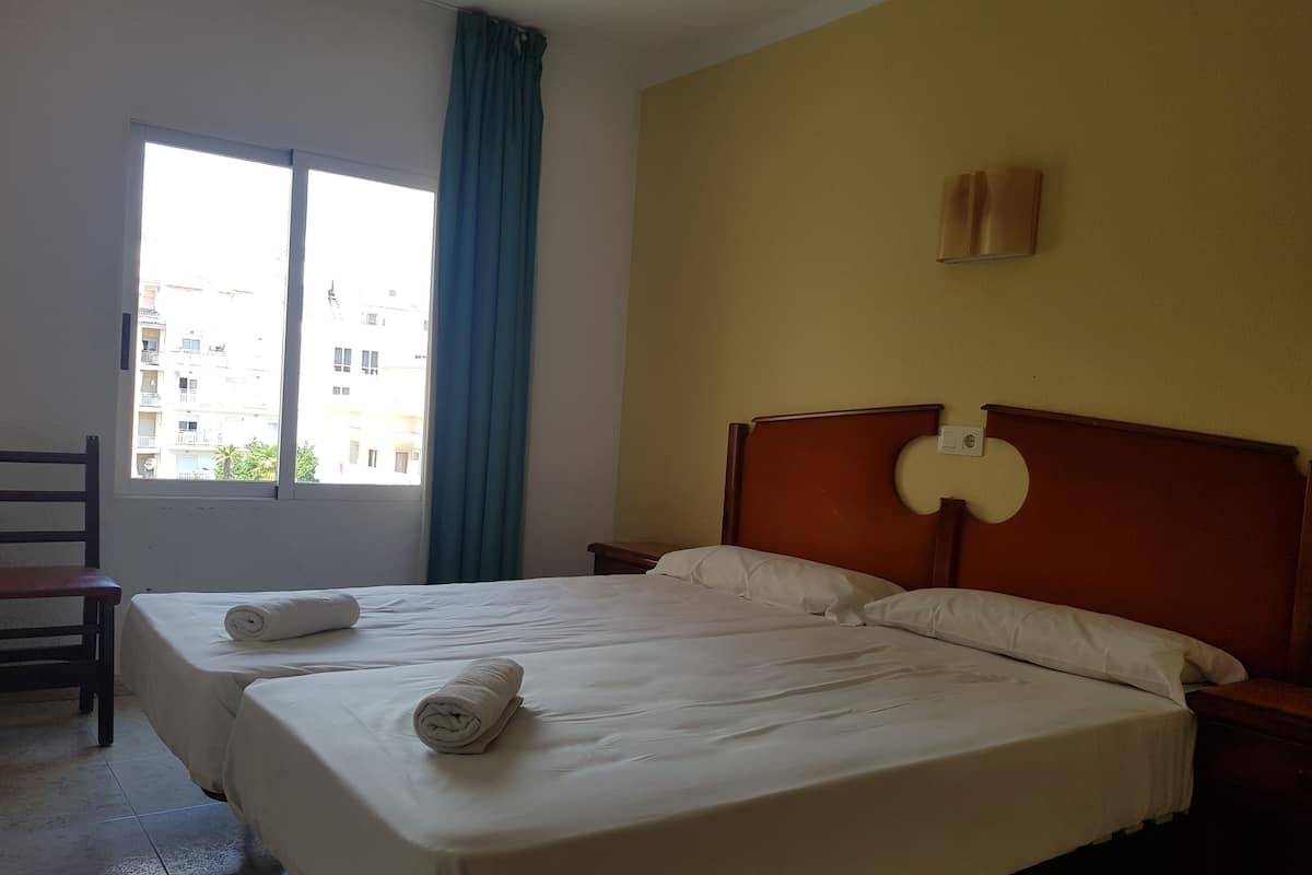 A hotel room with a large bed, two pillows, a wooden headboard, a nightstand, a chair, a window with curtains, and a view of buildings outside.