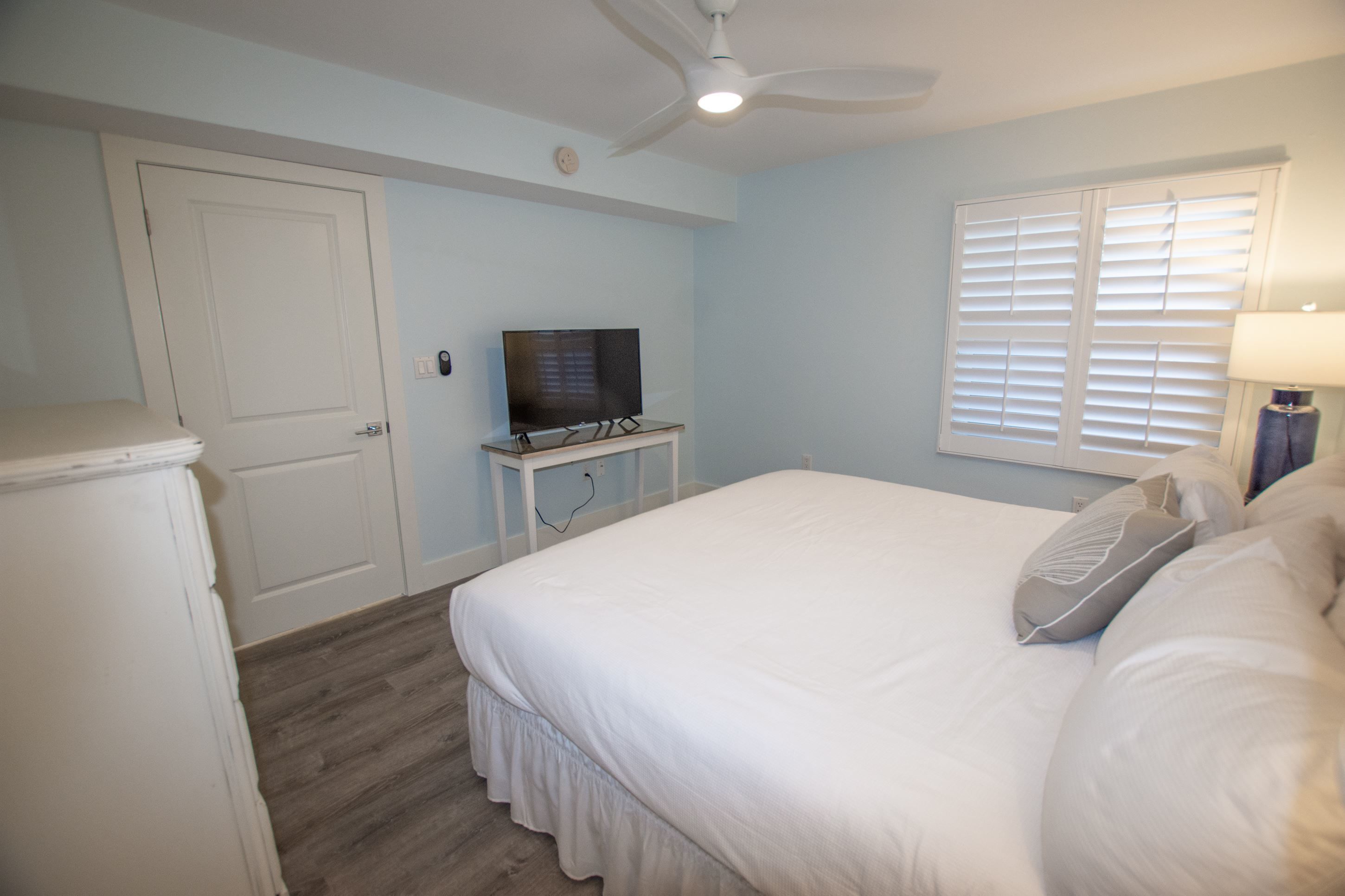 A bedroom with a bed, a television on a stand, a ceiling fan, a window with blinds, and a lamp on a bedside table.